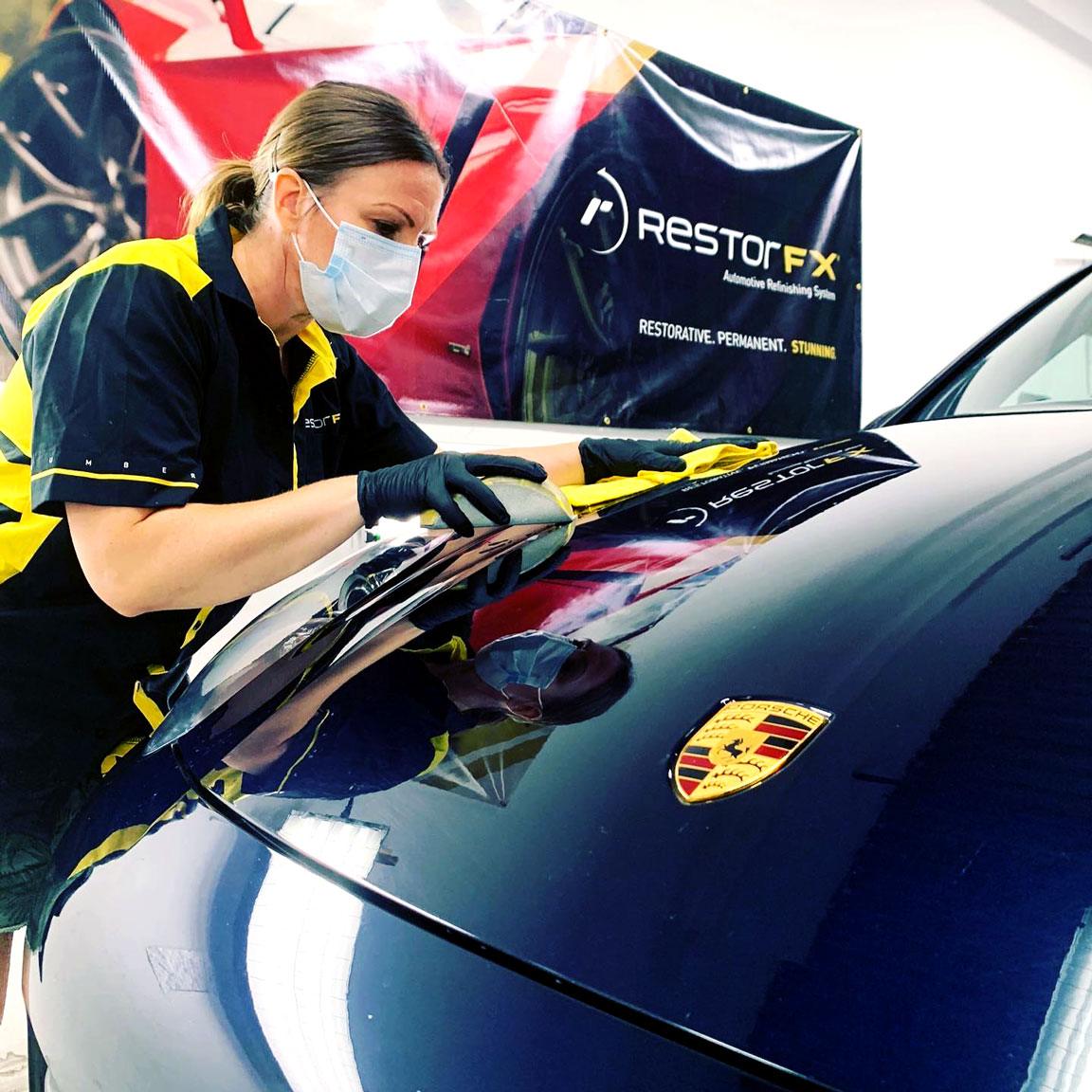 Technician wears mask while wiping the front of black Porsche during paint restoration at shop area