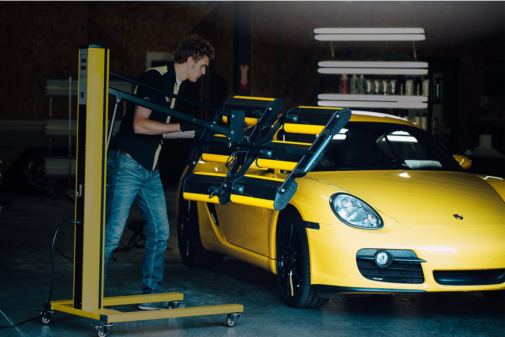 A RestorFX technician aiming an infrared lamp on a yellow sports car during restoration into a bright, shiny surface